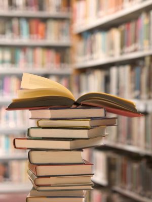 A stack of books line a table, with other books in the background