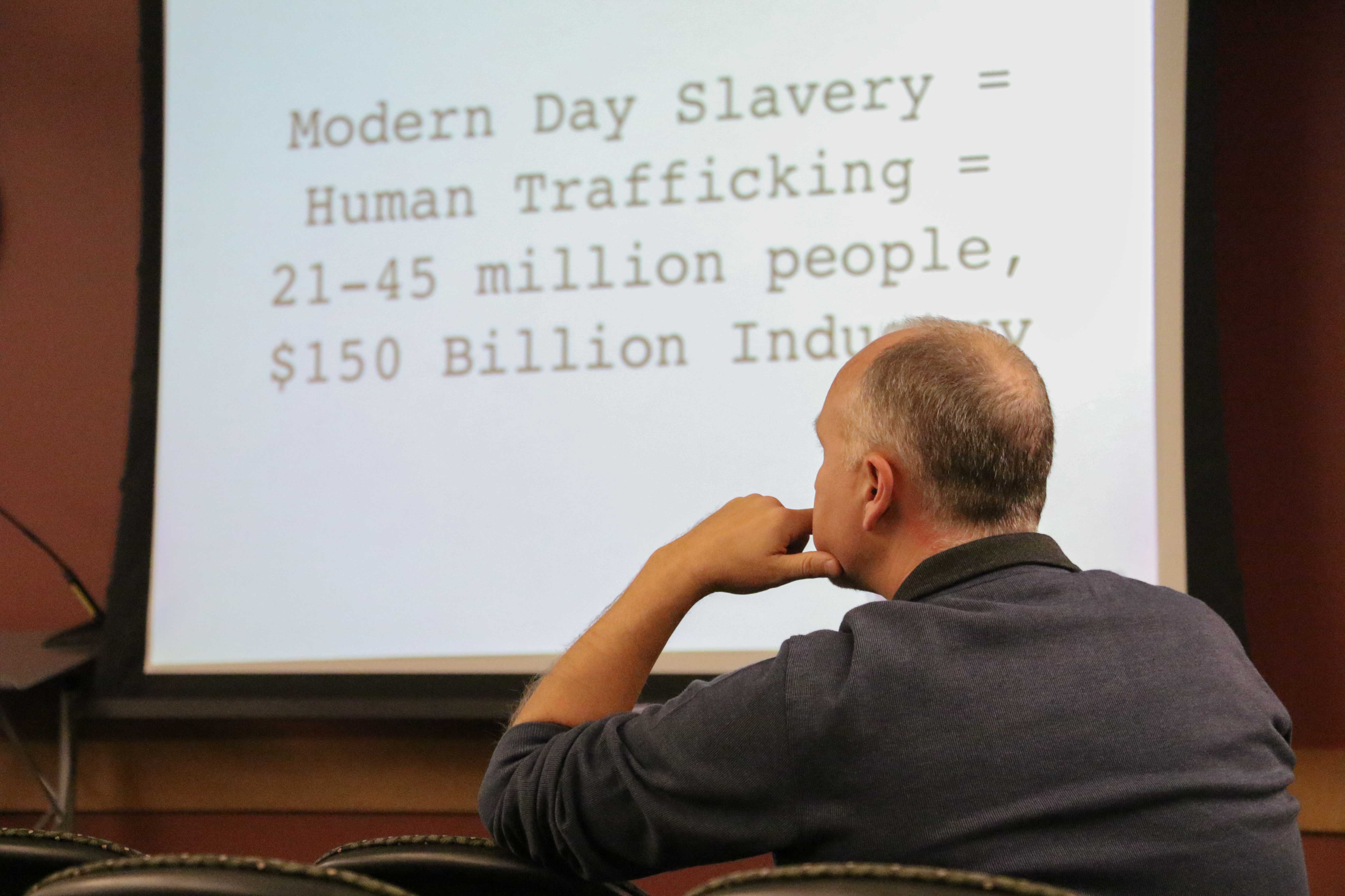 A man sits in front of a presentation on human trafficking. The slide behind him says 'Modern day slavery = Human trafficking = 21=45 million people, $150 billion industry.'