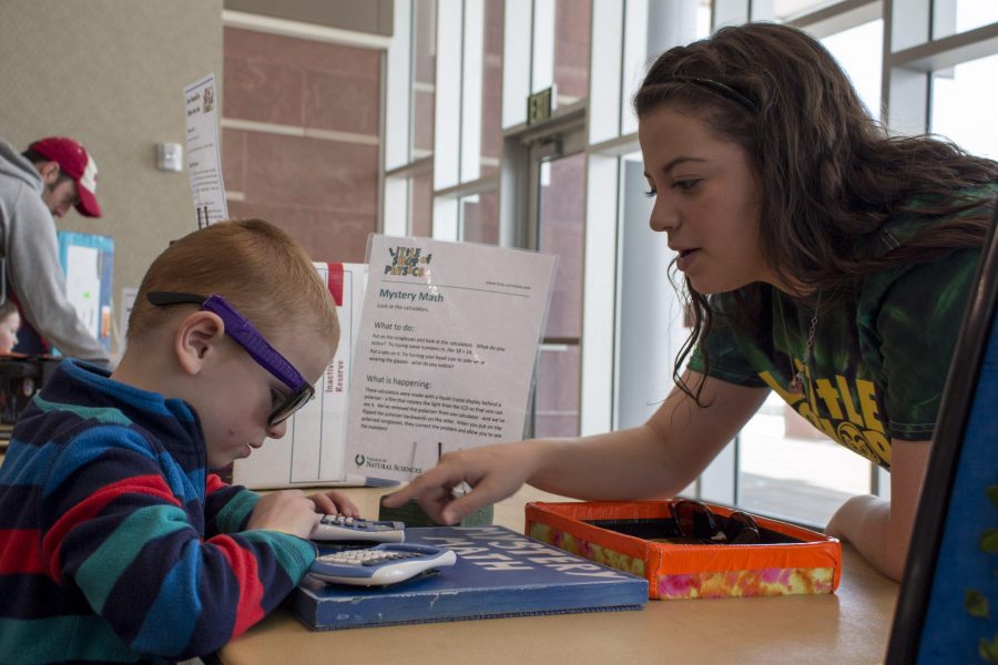 Kenzie Isbell, a volunteer for a Little Shop of Physics event, teaches a young boy the trick behind one of events stations. Photo credit: Brooke Buchan