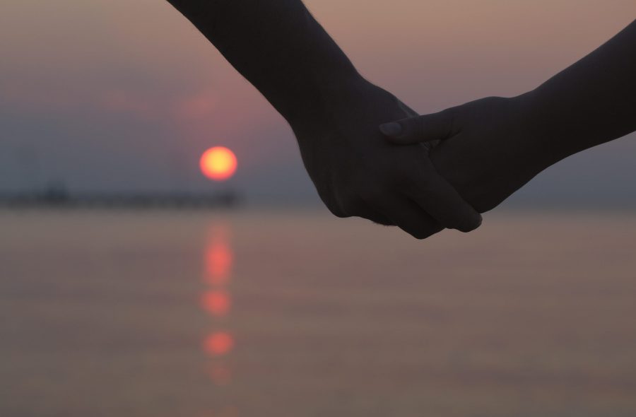 Close up of the hands of a romantic couple holding hands at sunset silhouetted against the colourful night sky with the orb of the sun visible below