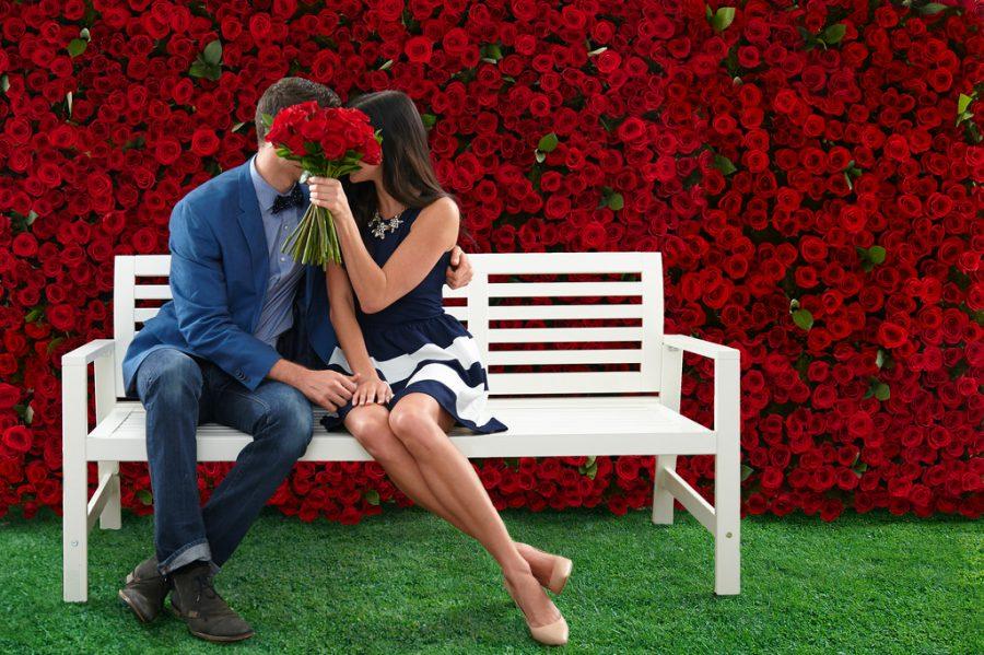 a man and woman kissing on a bench with their faces conceiled by a bouquet of roses.