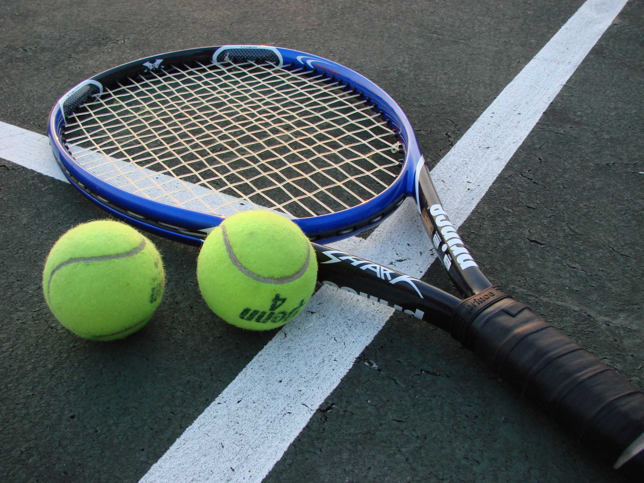 Two tennis balls sit to the left of a tennis racket on a tennis court.