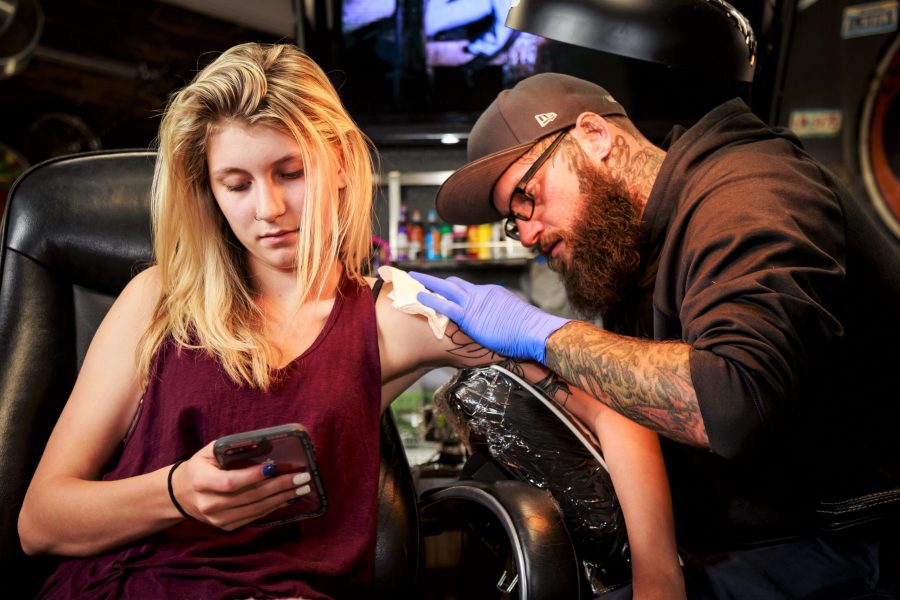 Lauren Farrer scrolls through her phone while tattoo artists Matt Phillips works to fill in a rose bouquet on her shoulder. Farrers first visit was to complete the outline work for the bouquet, but now that its healed, Phillips works to add the finishing touches to the piece. (Davis Bonner | Collegian)