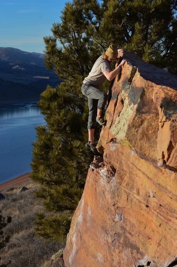 A rock climber clings to the edge of a small cliff with a view of trees, mountains and a lake in the background.