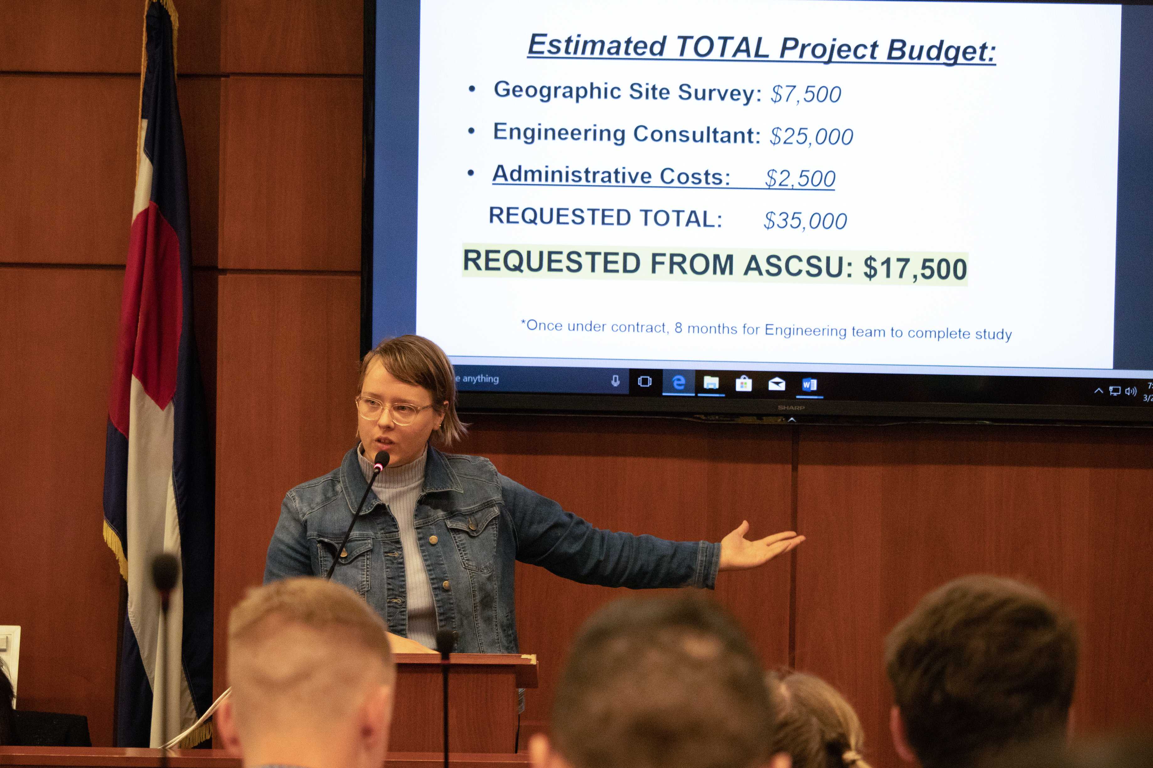 woman stands in front of powerpoint that says: "Estimated Total Project Budget: Geographic Site survey: $7,500; Engineering consultant: $25,000; Administrative costs: $2,500; Requested total: $35,000; Requested from ASCSU: $17,500