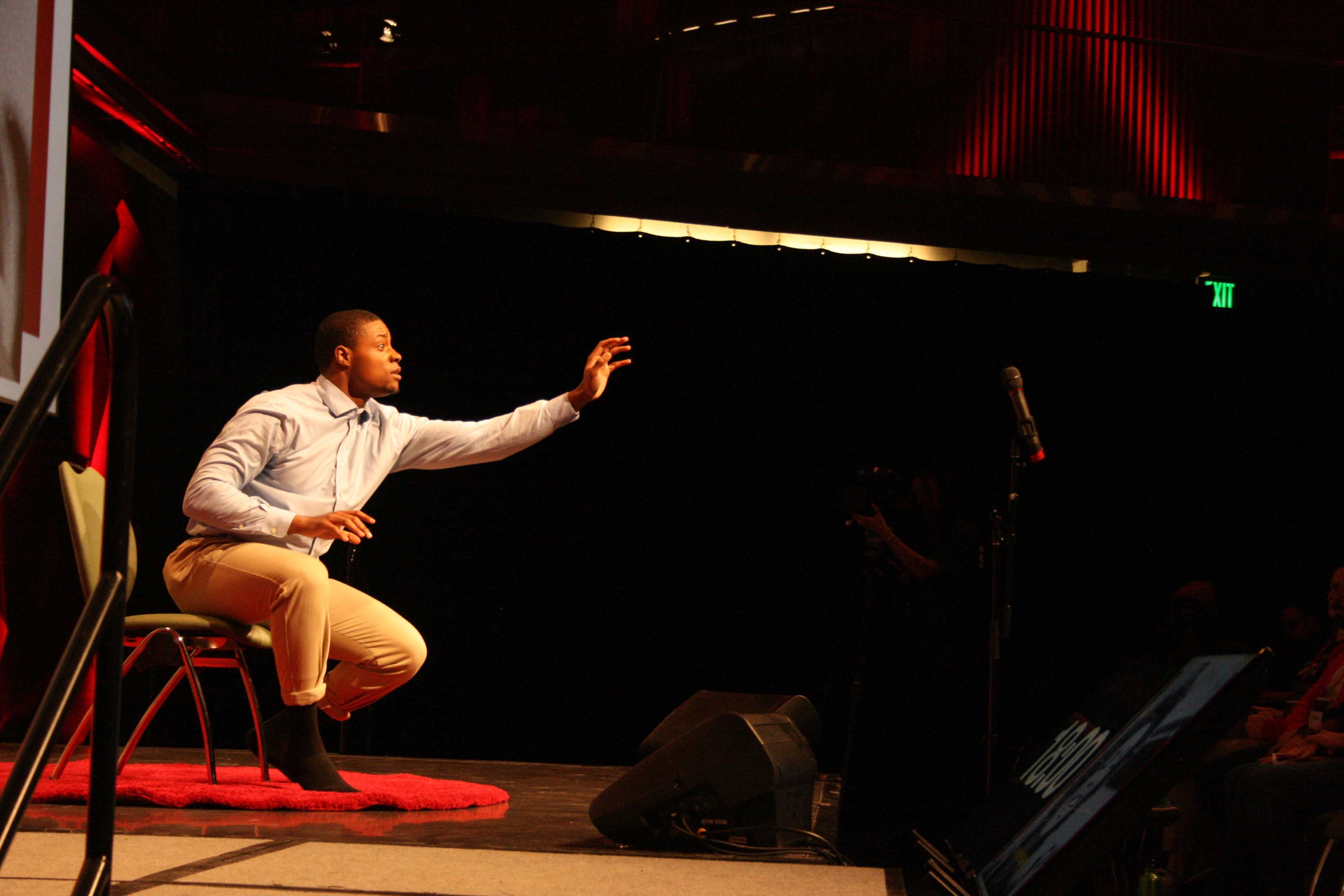 Young man sits on stage with an outstretched arm.