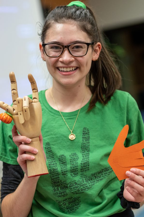 Junior Liberal Arts student Lu Epstein holds up wooden hand and paper heart both signing, "I love you," during the Smile event at the International House Thursday, Oct. 24. The paper hands are intended to be pinned to shirts in order to show solidarity with speakers of sign language. (Brooke Buchan | The Collegian).