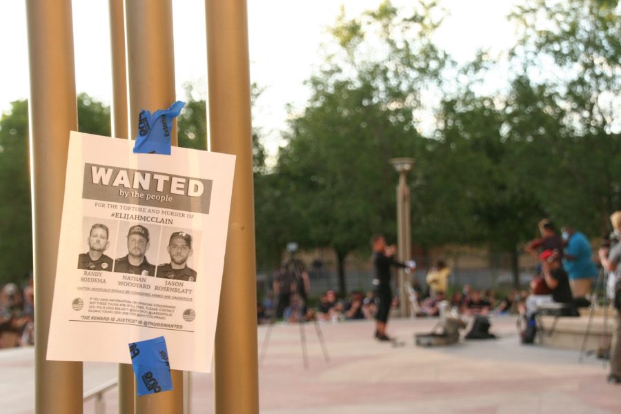 This sign was posted around the Aurora Municipal Center on June 27, calling for the arrest of the officers involved in the killing of Elijah McClain. Musicians play in his honor in the background. (Katrina Leibee | The Collegian)
