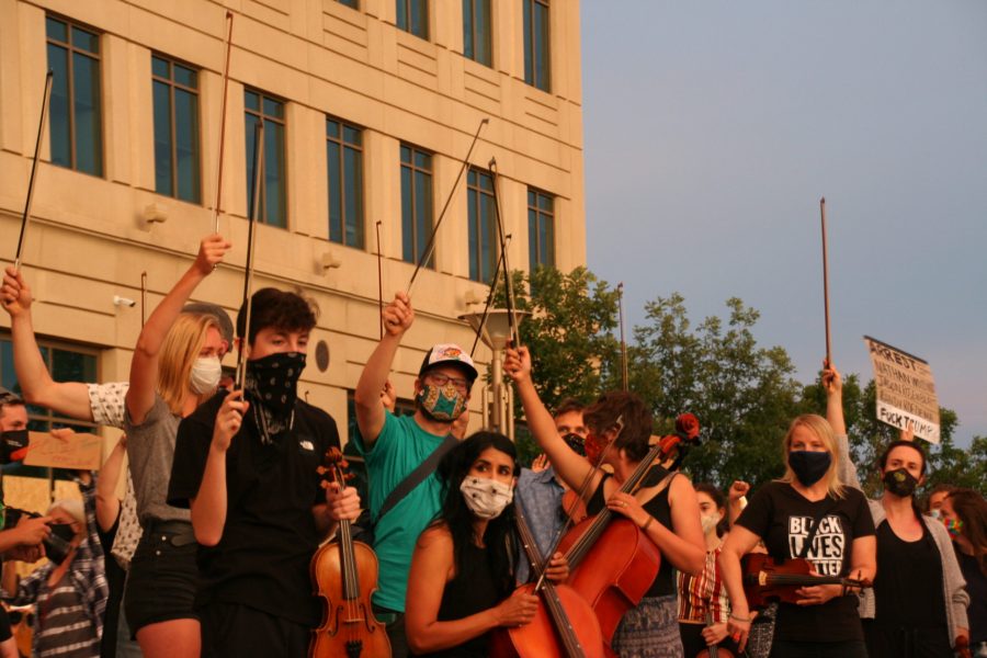 Musicians hold up their bows in honor of Elijah McClain while police officers push protestors out of the park. People linked arms to barricade the stage they were standing on and protect the musicians. (Katrina Leibee | The Collegian)