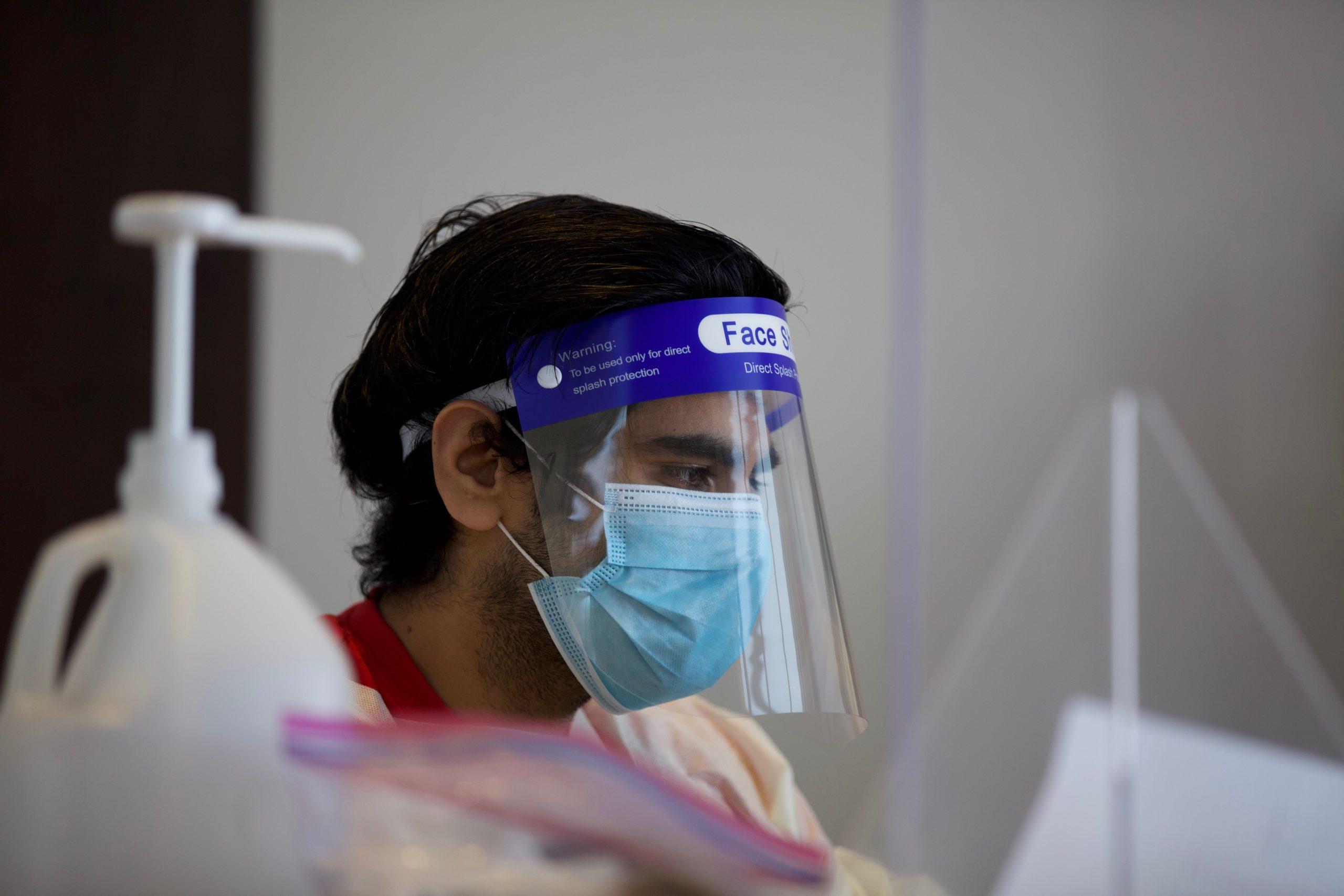 person with a mask and face shield sits at a desk