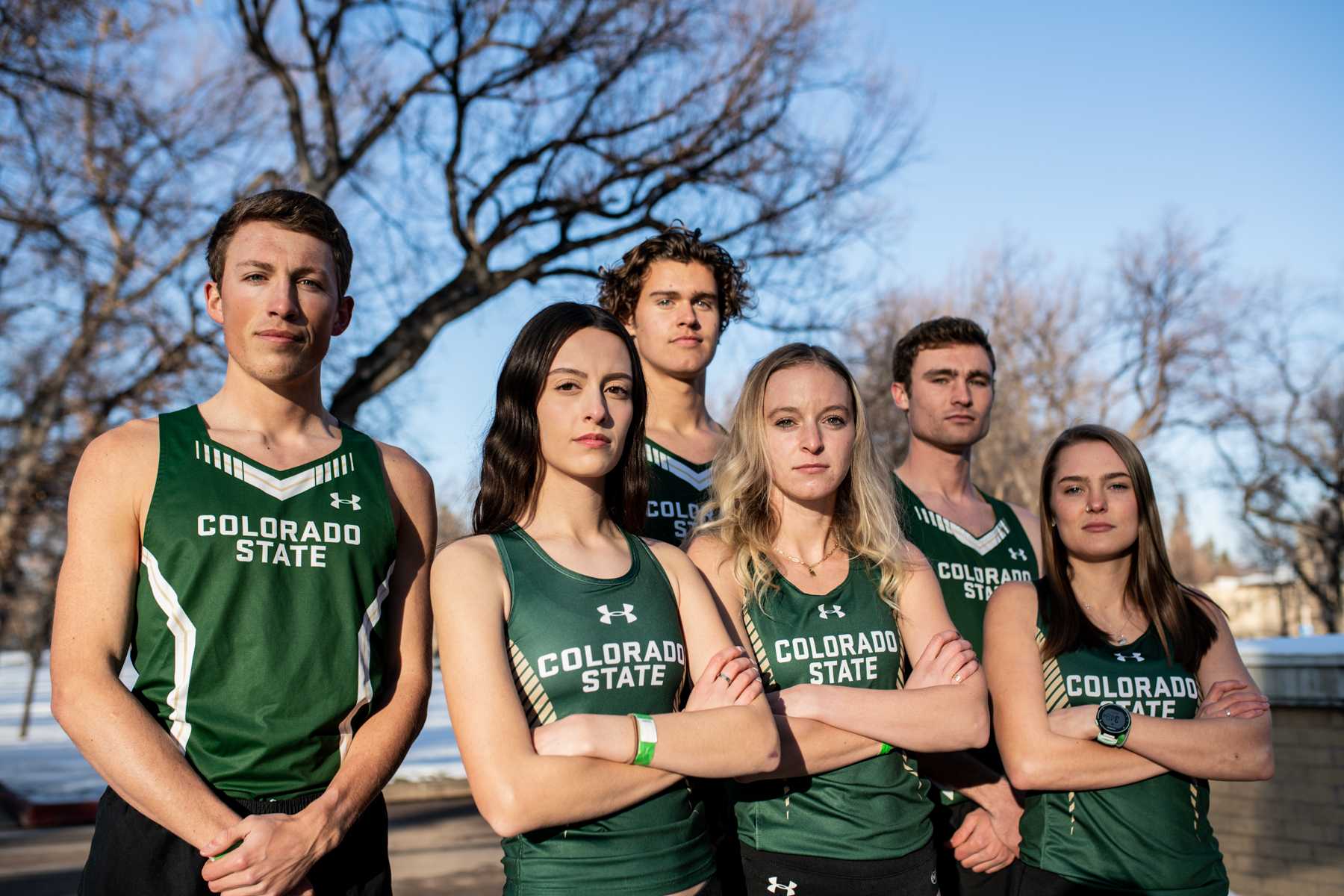 cross country team stands outside in green jerseys with arms crossed and trees in background