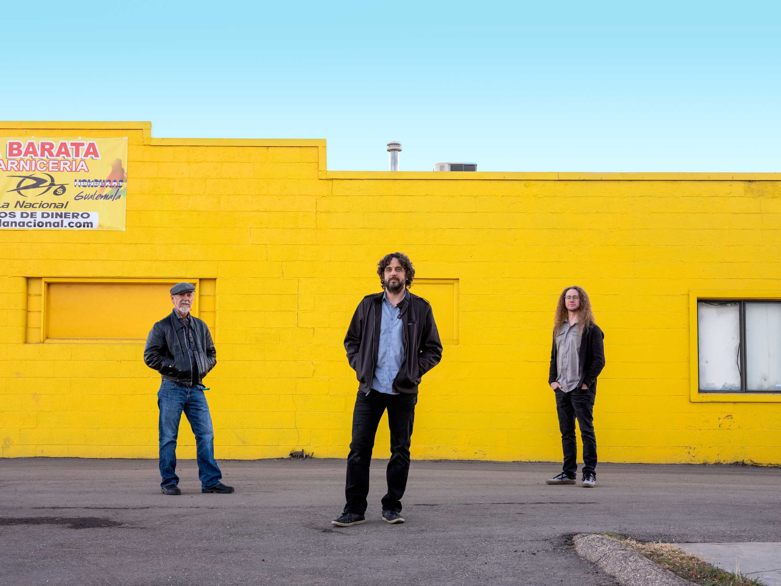 Three masculine people standing in a triangle formation against a yellow brick building. The sky is clear and they're standing on a road.