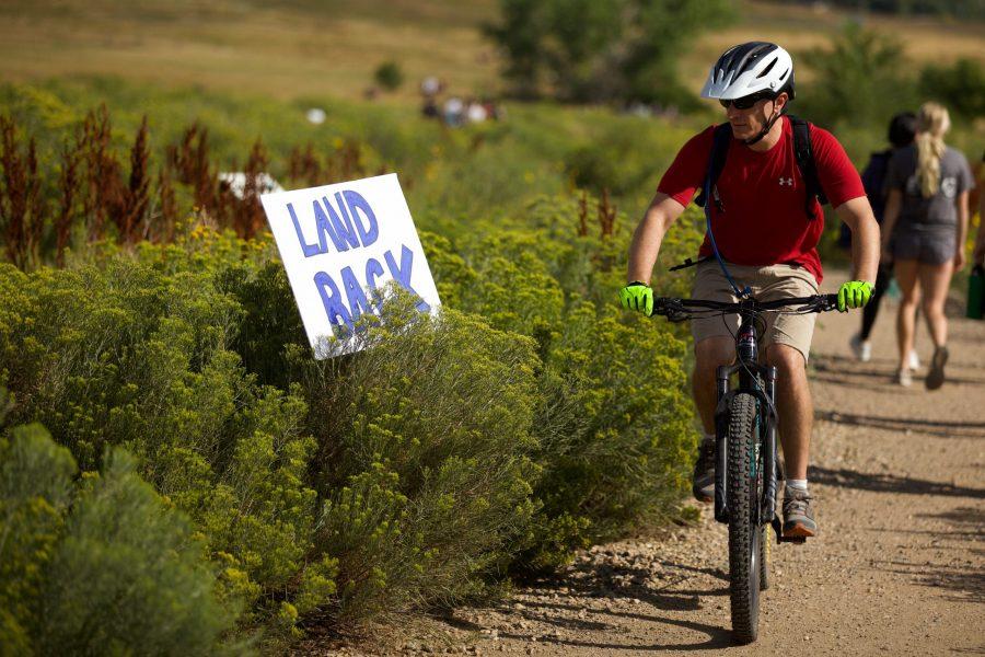 A biker at the Maxwell Natural Area Aug. 22 passes signs placed by the Intertribal Alliance for Right Relations. Demonstrators and activists were at the trailhead to explain the history of the Hughes land and their hope for it to be used as a gathering place for Indiginous people. (Ryan Schmidt | The Collegian)