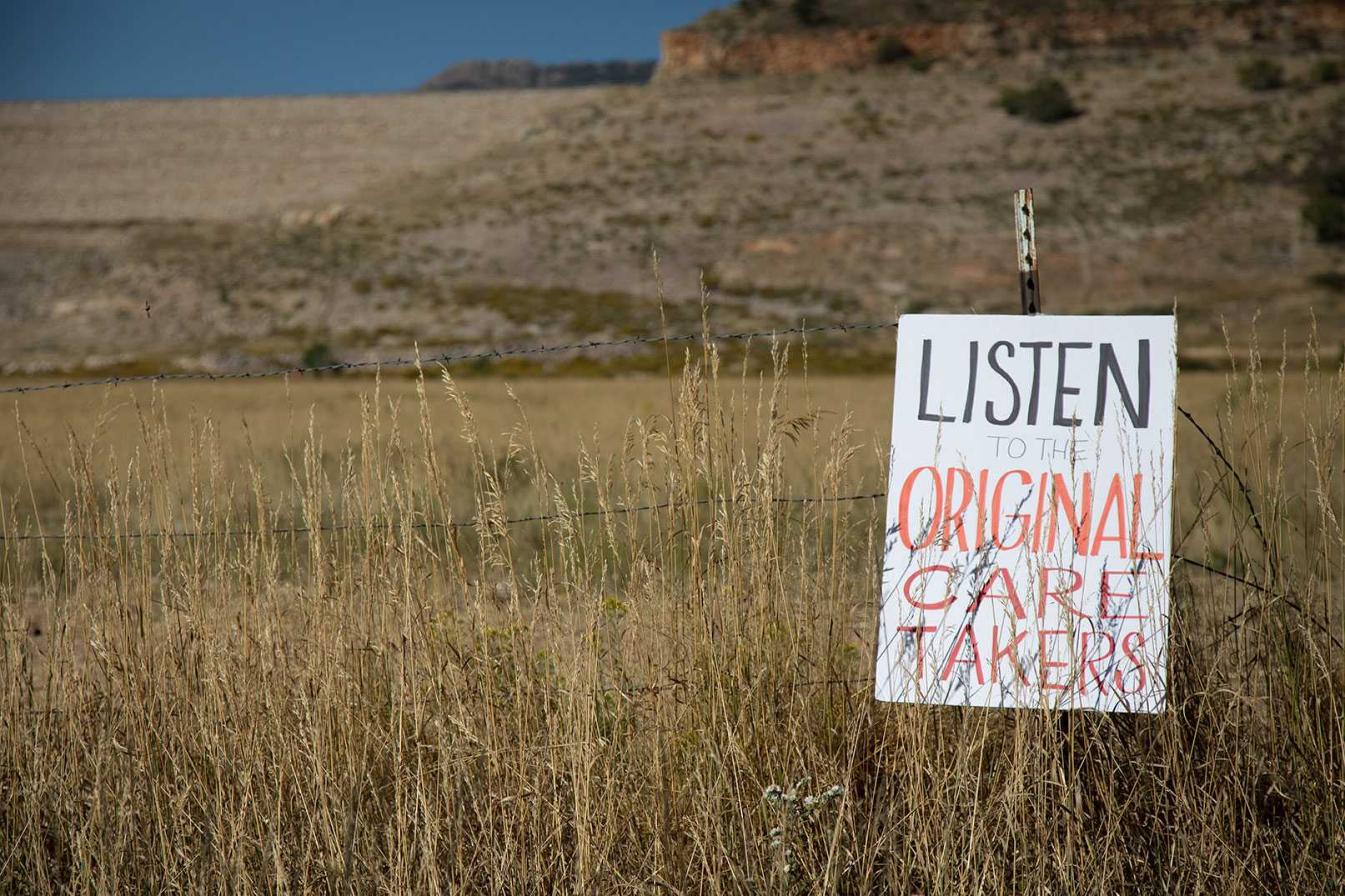 A sign that says "Listen to the origianl caretakers" is proped up along a barbed wire fence. The foothills stand just behind the sign.