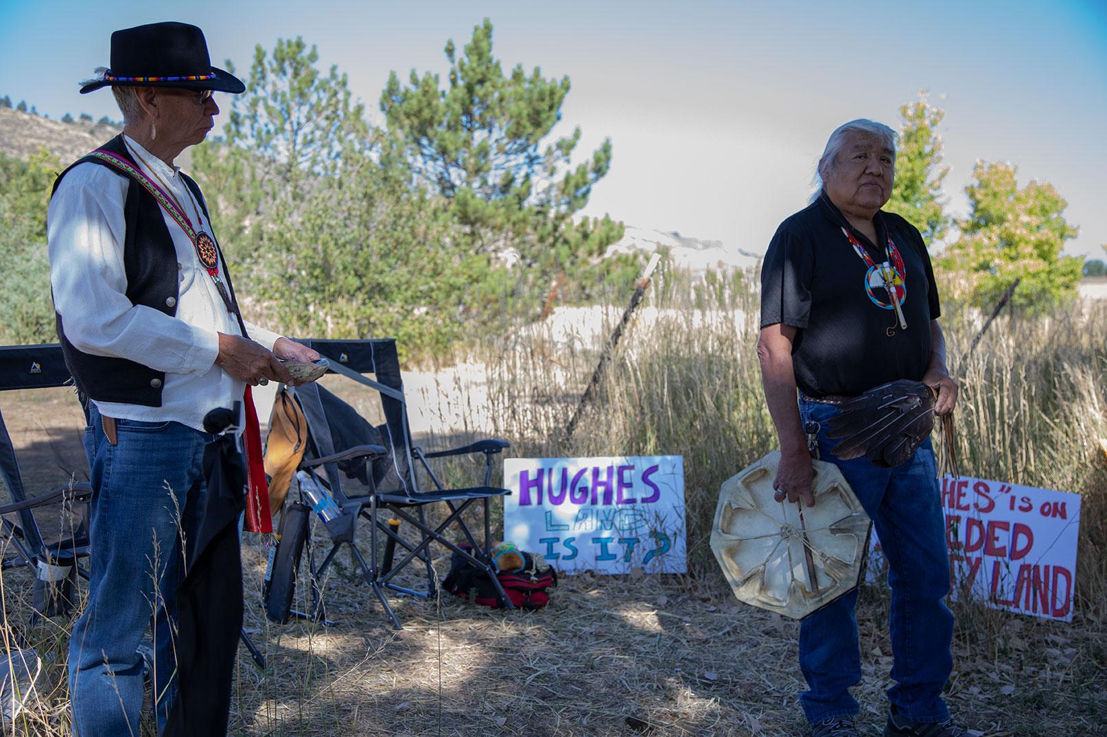 Two Indigenous men stand in the shade of trees, speaking to a group of people.