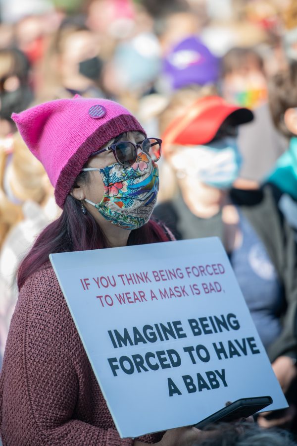 A march-goer wearing a version of the iconic pussyhat watches speakers on the steps to the Colorado State Capitol while holding a sign comparing mask mandates to restrictive abortion laws Oct. 2. (Lucy Morantz | The Collegian)