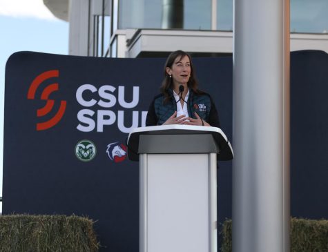 A woman gestures by putting her hands together while smiling and standing behind a silver podium. In the background is a sign that reads "CSU Spur."