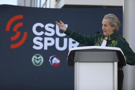 A woman gestures with her right hand while standing behind a podium. In the background of the photo is a sign that reads "CSU Spur."
