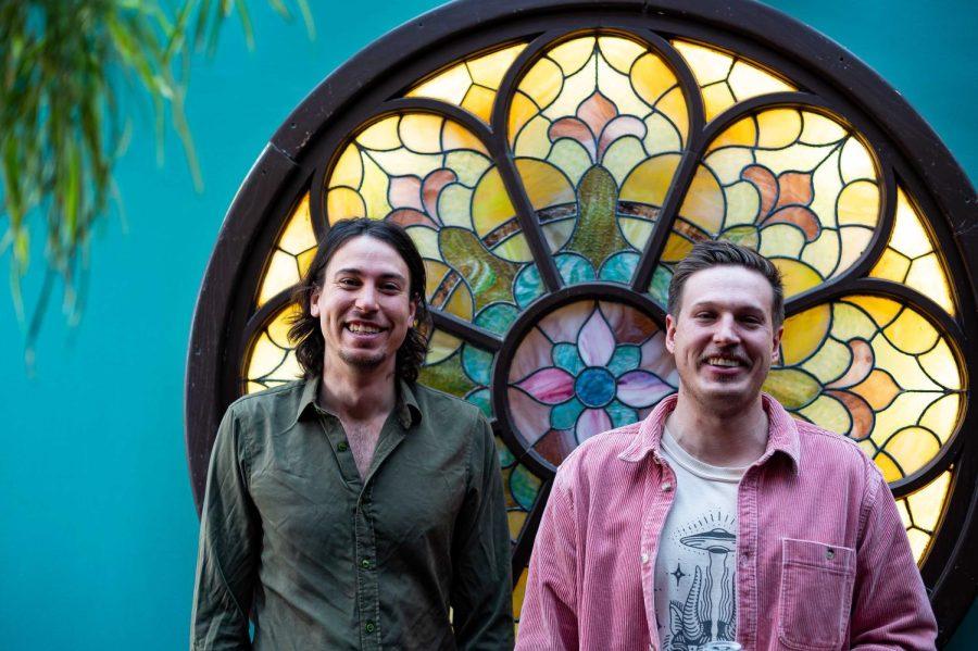 Two men stand in front of a colorful stained glass mandala.