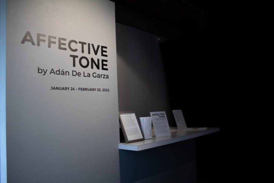 A gray wall features the text Affective Tone by Adán De La Garza, January 24 - February 25, 2022. This gray wall is on the left of a black wall and a counter with various pamphlets.