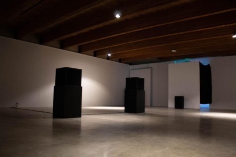 A dark room with white walls and cement flooring features black stereos. A green exit sign is seen peaking over the wall.