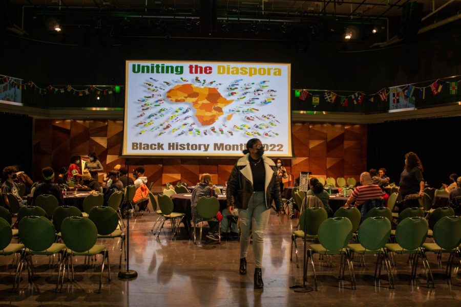 Community members gather in small tables in a dark theater, which has a projector screen in the middle. The screen says Uniting the Diaspora and Black History Month 2022. The screen has a picture of the African continent in the center.