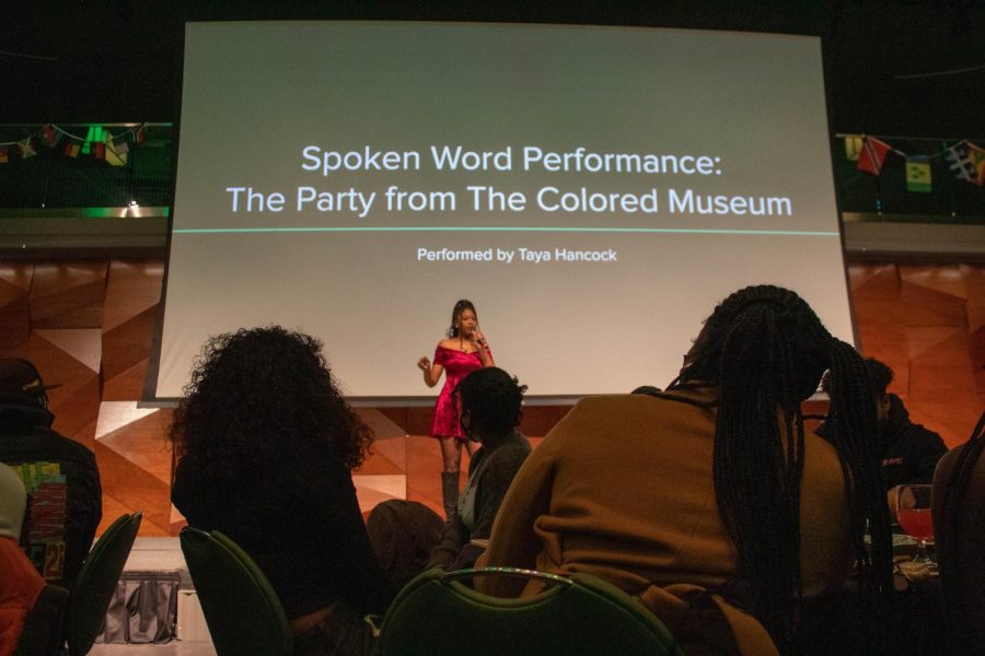 Taya Hancock performs on a stage, wearing a mid-thigh length red dress and braids. She performs in a theater with brown geometric walls and a projector is behind her. The projection says Spoken Word Performance: The Party from The Colored Museum, Performed by Taya Hancock.