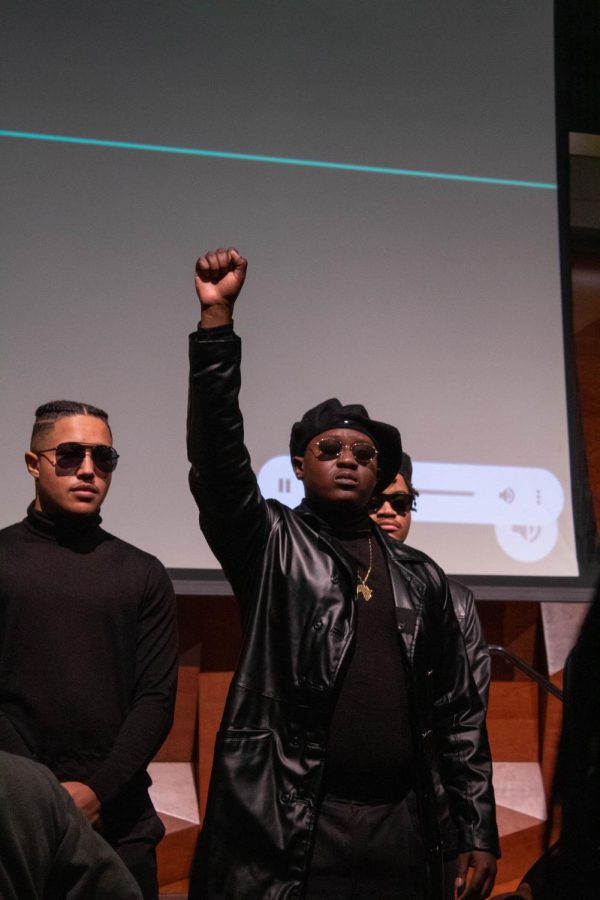 Joseph Adenowa, who is dressed in all Black to represent civil rights activist Fred Hamptom, stands on stage beside two other men. All of them are wearing sunglasses, and a projector is behind them on the stage they stand on.