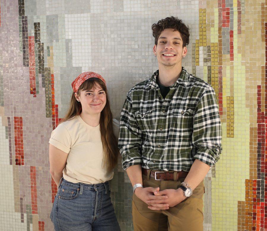 Two people stand in front of a colorful wall and smile at the camera