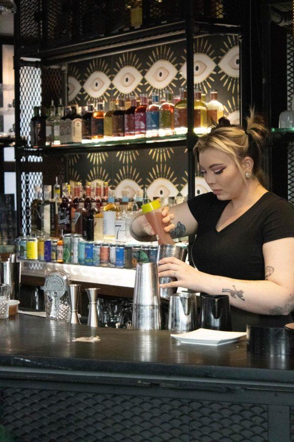 A woman with dyed hair mixes a drink behind a bar. Behind her is a mix of non-alcoholic beverages and a wallpaper with an eyeball pattern.