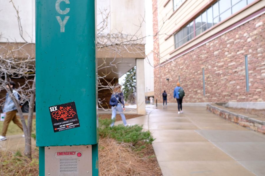 Students walk by a sticker on an emergency call box to the north outside Albert C. Yates Hall March 29.