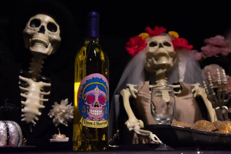 Two skeletons dressed in bride and groom wedding attire sit at a table with fake fruit and a bottle of wine for Dia de los Muertos at Global Village Museum of Arts and Cultures