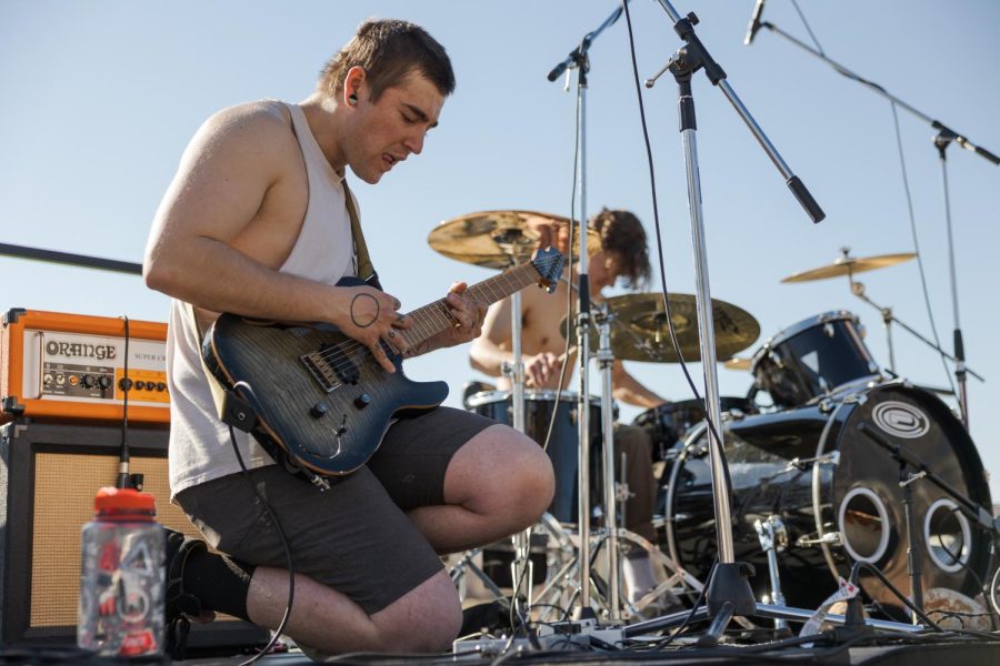 A man kneels while playing guitar in front of a man playing drums.