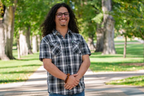 A man in a plaid shirt smiles at the camera with his hands clasped in front of him, standing in front of a background of trees and grass.