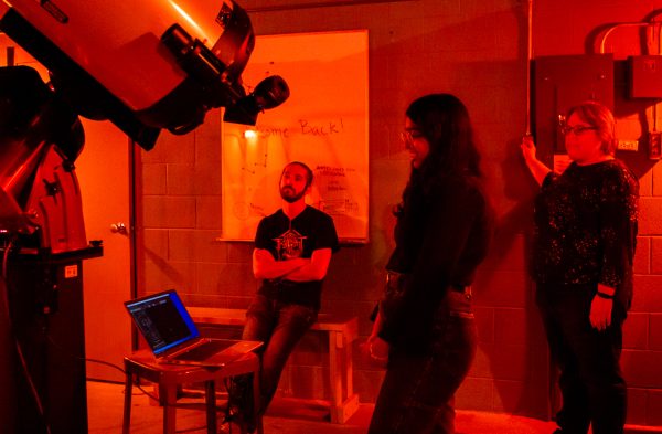 People observe and discuss the stars around a large viewing telescope. The room glows dimly red.