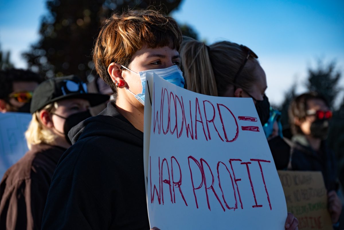 A protester stands with a sign and listens to a speaker talking about their experiences at the "Die-In at Woodward" protest.