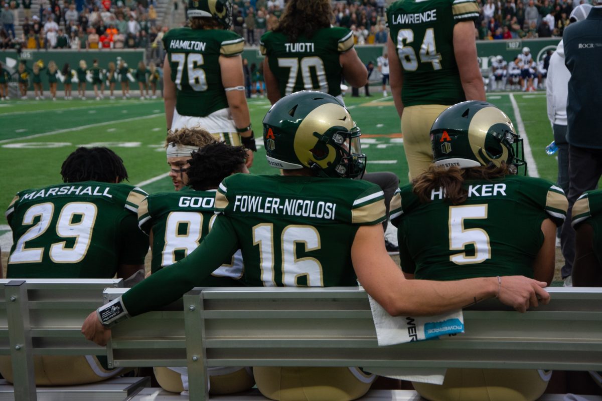 Viewed from behind, Colorado State University player in green, gold and white uniform sits on the sideline bench with other players during a game.