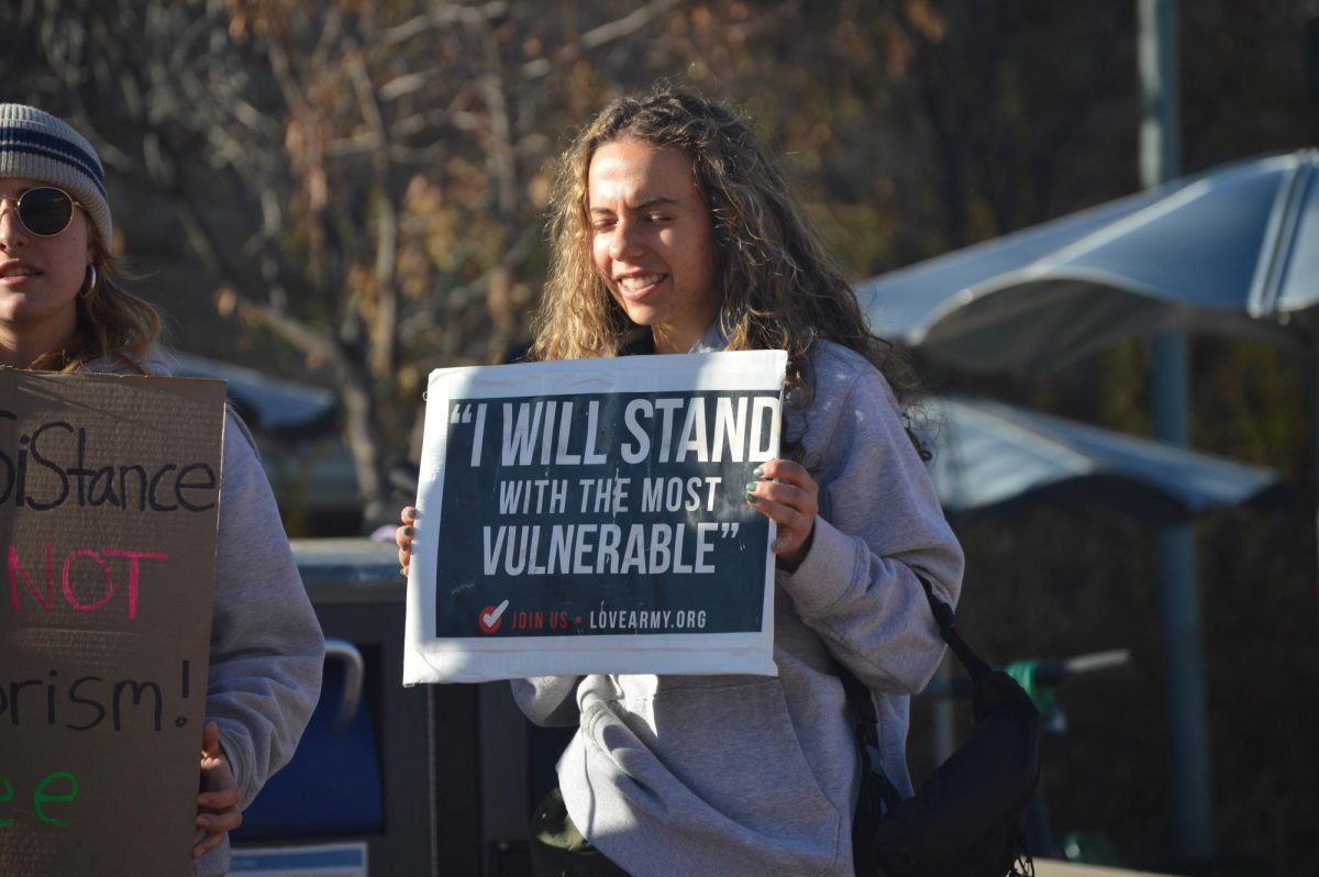 A smiling protester holds a sign reading, I will stand with the most vulnerable.