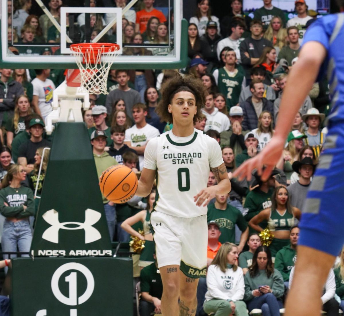 A male basketball player weraing a white and green jersey with the number 0 dribbles the ball down the court as a packed crowd watches him.