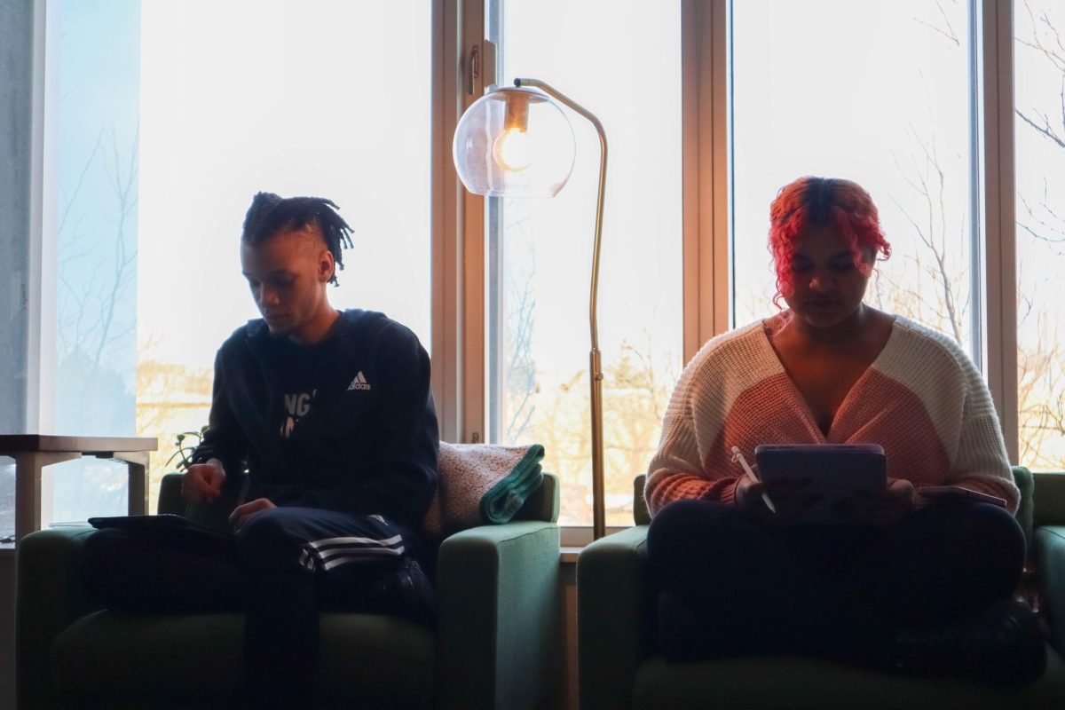 Two students sit in couches side by side while looking down at their tablets.