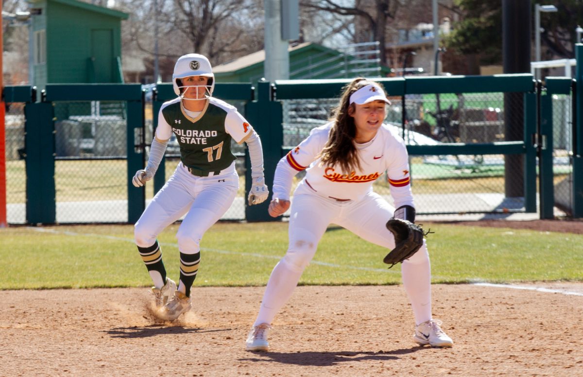 A woman wearing a green and white uniform runs behind her opponent, a woman in a red and white uniform, during a softball game.