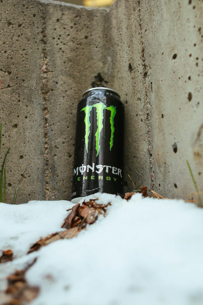 A Coca-Cola Monster Energy drink sits upright in the snow against a concrete wall. March 26.