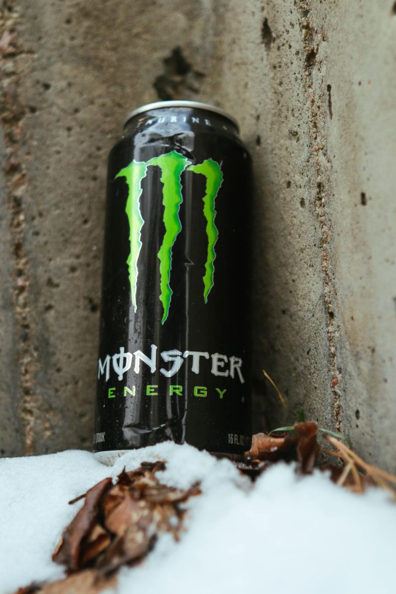 A Coca-Cola Monster Energy drink sits upright in the snow against a concrete wall. March 26.