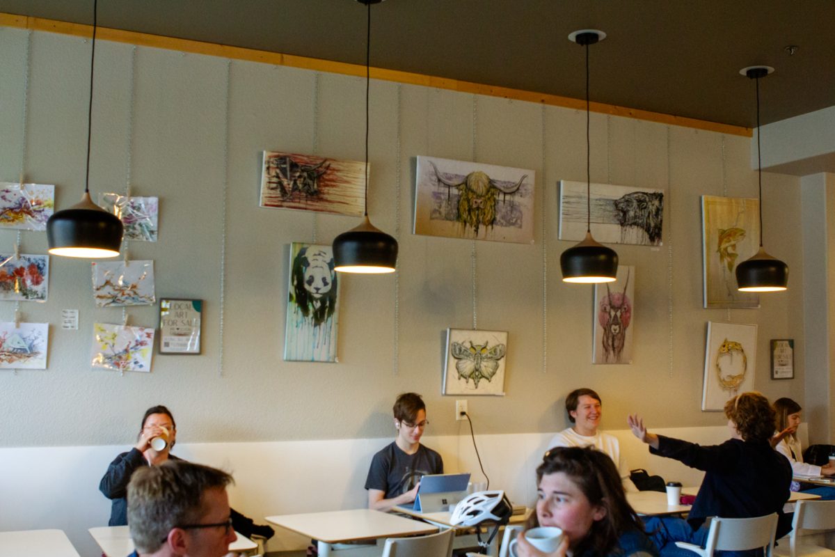 Art hangs on a wall. Customers chat with one another at tables against the wall underneath.