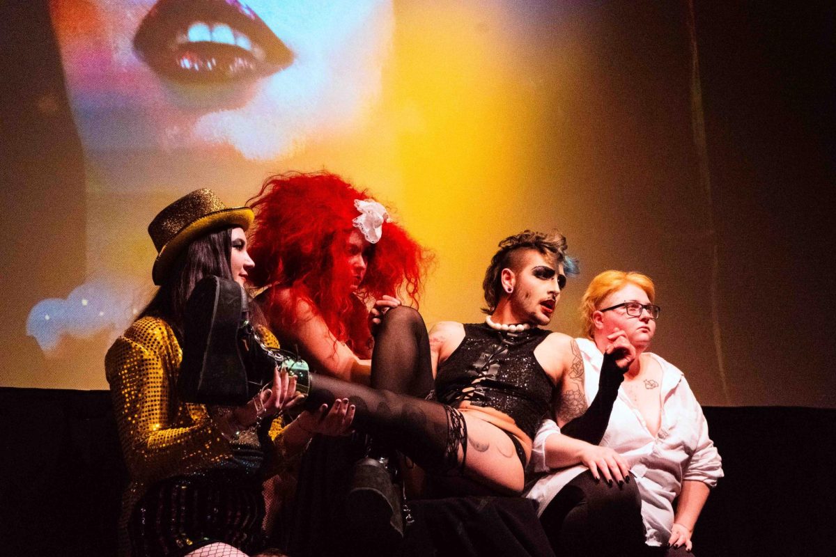 Three performers sit with a fourth performer wearing a pearl necklace, black tank top, black garters and black boots sprawled across their laps.