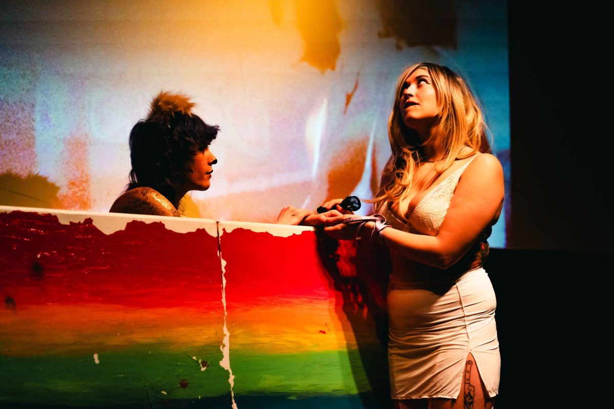 One performer wearing a lion costume sits behind a piece of wood painted with an ombre rainbow. Another performer wearing a tight white dress stands to the right, looking up.