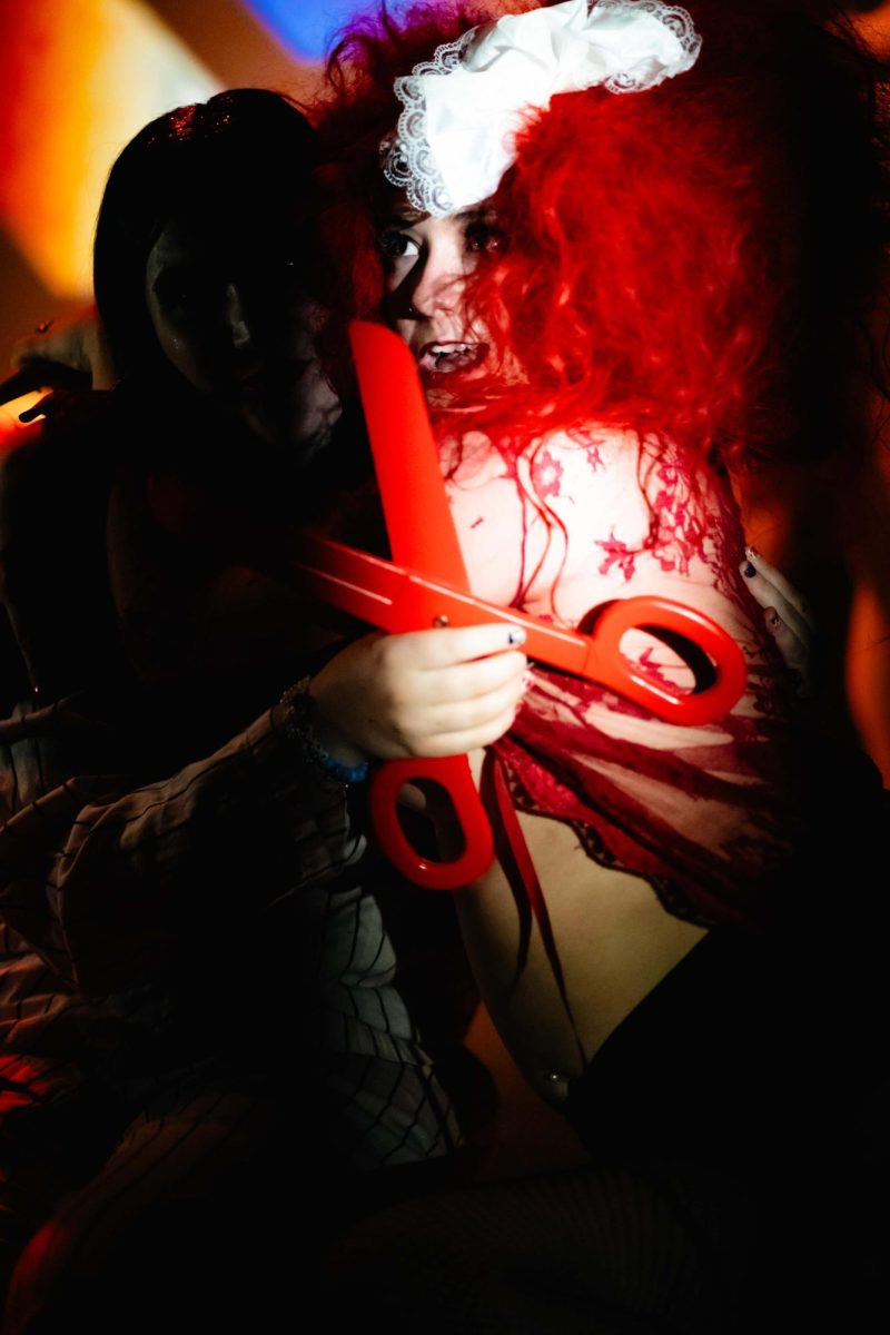 Two performers embrace, one in darkness and holding a comically large pair of red toy scissors and the other wearing a large red wig and gasping.