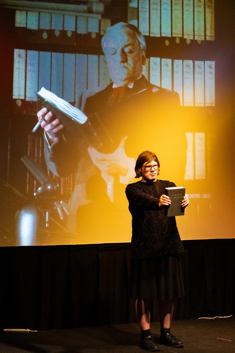 A performer in a black dress holds a black book directly in front of them with their arms straight out. The background is a projection of an old man in a suit reading from a large book.