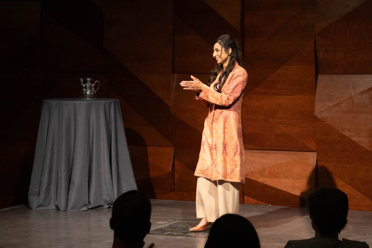 A woman standing on a stage looks to her right with her hands pressed together in front of her.