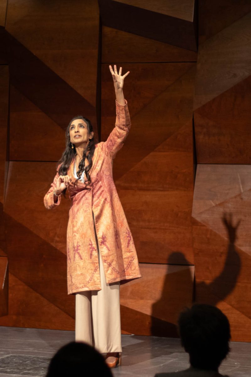 A woman speaks on a stage with one hand held above her head.